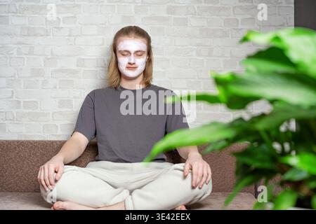 Relaxed young man closing eyes and sitting in metitation pose with cosmetic clay mask on his face. Relaxing, balanced lifestyle, home beauty treatment Stock Photo