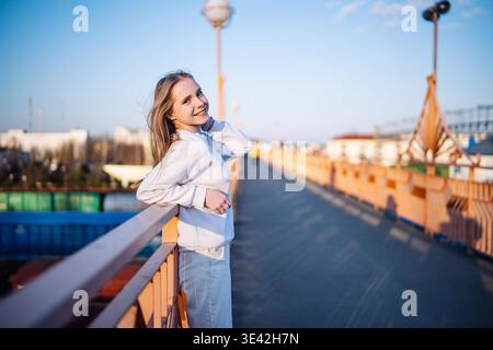 Smiling young woman leaning on a bridge railing during a sunny day Stock Photo