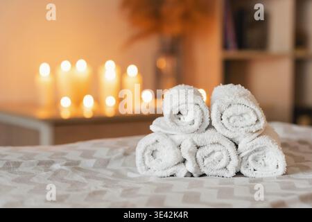 Rolled white towels stacked on a bed or massage table, offering a concept of spa, wellness, and relaxation with blurred candles Stock Photo