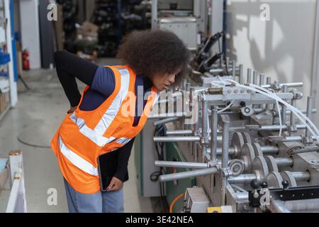 African American female industrial worker having physical workload and back pain while checking machinery, workplace injury in manufacturing Stock Photo