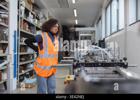 African American female industrial worker having physical workload and back pain while checking machinery, workplace injury in manufacturing Stock Photo