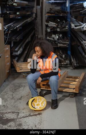 Fatigued African American female warehouse worker taking off a yellow hard hat and sitting down to rest after a hard workday. Stock Photo