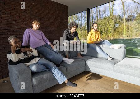 Four friends lounging on a sofa in a bright, modern living room with large windows and natural light Stock Photo