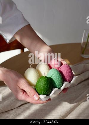 Female hands holding a ceramic tray with colorful beeswax candles in the shape of decorated Easter eggs. Spring holiday home decor. Close-up. Stock Photo