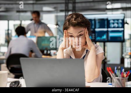 Frustrated female office worker sits at desk, feeling stressed and burned out while working on laptop. Caucasian woman with hands on head, showing signs of exhaustion and tension in busy workplace. Stock Photo