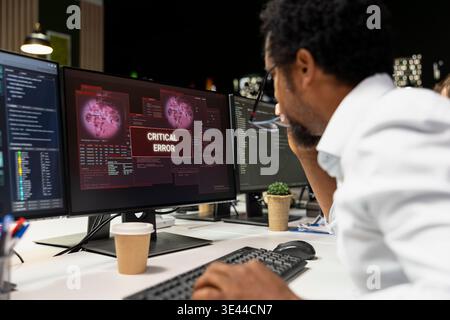 Technician at office desk thinking how to fix critical error warning displayed on PC monitor. African american man in tech startup brainstorming how to fix computer bug affecting server network Stock Photo