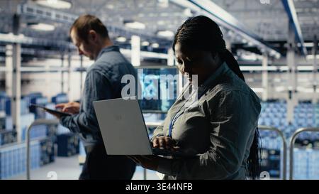 Data center technician deploys machine learning software on infrastructure racks. African american woman in server farm trains AI systems with datasets using laptop to support applications, camera B Stock Photo