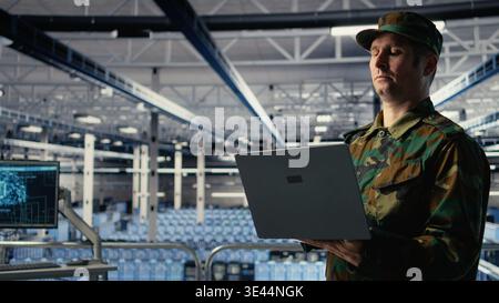Soldier in server hub using laptop to deploy AI tech, modernizing defense infrastructure. Military data center specialist integrating artificial intelligence technology using notebook, camera A Stock Photo