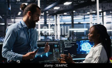Manager supervising woman in data center coding with generative AI assistance. Supervisor oversees work by african american IT expert leveraging artificial intelligence large language model, camera A Stock Photo