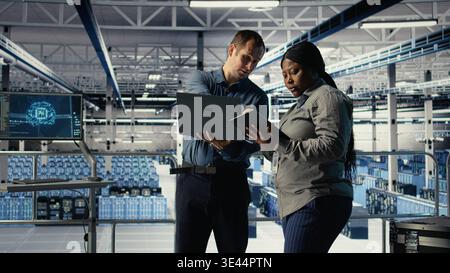 Team of data center programmers monitors deep learning process with laptop, reviewing operational metrics. Teamworking server hub colleagues review neural networks reports during inspection, camera A Stock Photo