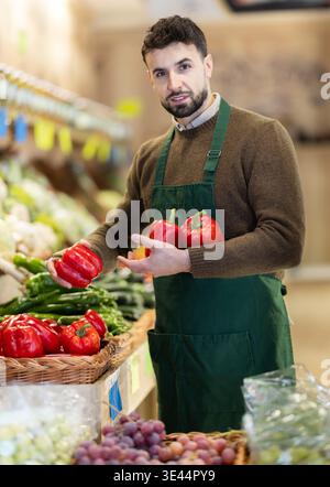 Young male seller putting red bell peppers on counter in grocery market Stock Photo