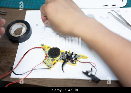 Close-up of hands repairing yellow electronic circuit board with gears and wires on white paper in Stock Photo