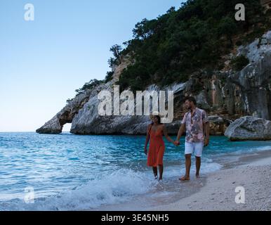A couple enjoys a romantic walk on the sandy shores of Sardinia, Italy, as gentle waves lap at their feet. The picturesque landscape and warm colors of the sunset create a perfect holiday atmosphere. Stock Photo