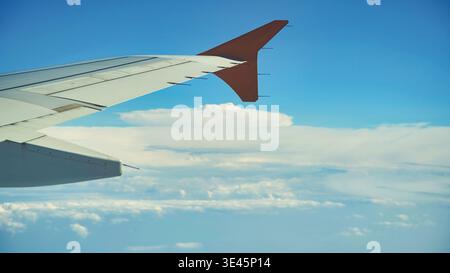 Red and white airplane wing soaring through blue sky, fluffy white clouds below evoking a sense of freedom and travel Stock Photo