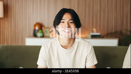 Comfortable, portrait and relax with happy man on sofa in home living room for weekend break as student. Confidence, me time and smile with Asian Stock Photo
