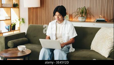 Laptop, relax and man on sofa in home with online application for university scholarship. Education, technology and Asian male student with computer Stock Photo
