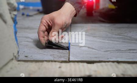 Close-up of a tiler's hand using a leveling system to install gray ceramic floor tiles. Focus on the wedge and clip tool. Concept of home improvement, Stock Photo