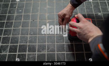 Close up, high angle view of a craftsman's hands using a red laser level tool. The worker is measuring on a dark gray tiled floor during renovation. F Stock Photo