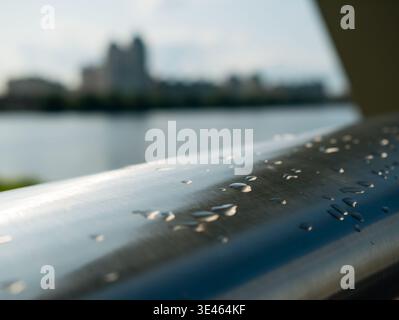 Close-up of rain droplets on stainless steel bridge railing above a wide river illuminated by sunlight after rain. Shiny metal texture with water drop Stock Photo