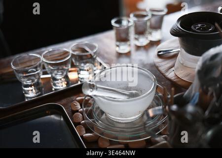 Clear glass cup filled with steaming hot liquid sitting on wooden table beside small empty glasses and dark teapot setting calm relaxing and soothing Stock Photo