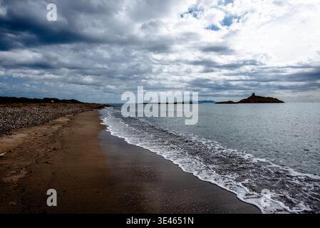 Quiet sandy beach with small waves rolling onto shore and a rocky island on the horizon under a dramatic cloudy sky. Stock Photo