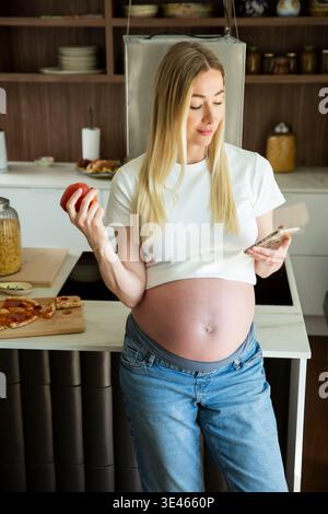 Pregnant woman choosing apple as healthy snack while using smartphone in kitchen. Maternity nutrition, digital health tracking and wellness concept. Stock Photo
