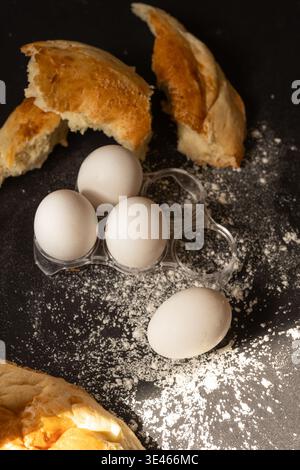 Composition of four white eggs in a clear plastic tray, one loose egg, and torn bread pieces on a dark surface dusted with flour. Stock Photo