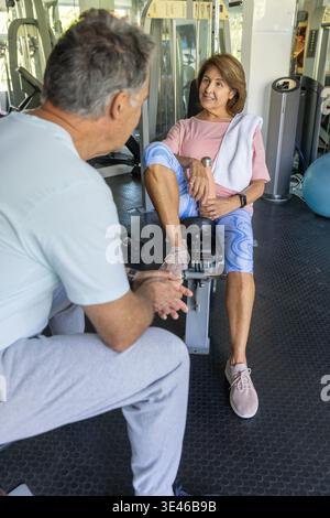 Senior woman and mature adult man sitting at gym using leg-extension bench with towel and dumbbells. Fitness, training, partners, equipment, weights, Stock Photo