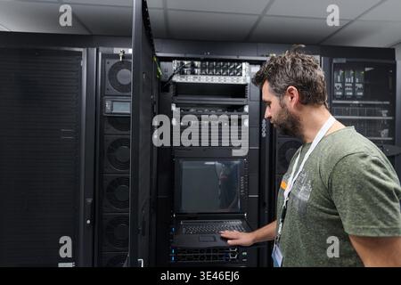 An IT technician inspects a server rack and rack-mounted console in a secure data center, using a laptop to perform maintenance, monitoring, and network troubleshooting with concentration and professionalism. || Model released Stock Photo