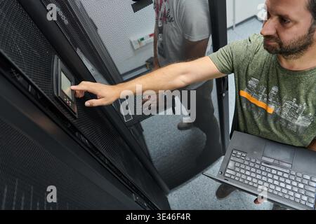 IT technician uses a touchscreen control panel on a server rack in a data center, holding a laptop while monitoring equipment and performing network or hardware diagnostics and maintenance. || Model released Stock Photo