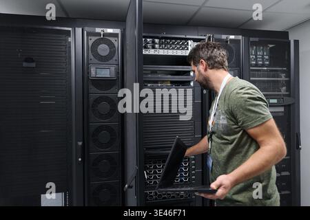 Male IT technician working in a data center server room inspecting rack-mounted servers with a laptop, performing maintenance, troubleshooting and network monitoring in a secure, climate-controlled facility. || Model released Stock Photo