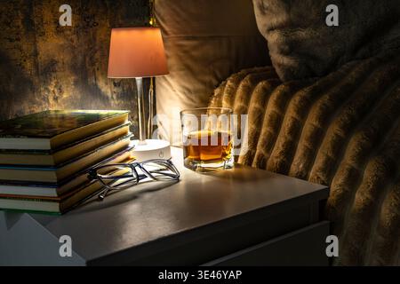Books placed on a nightstand in the bedroom, next to the books there is a night lamp that casts a warm light on the pages of the books, on the table t Stock Photo