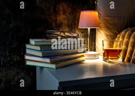 Books placed on a nightstand in the bedroom, next to the books there is a night lamp that casts a warm light on the pages of the books, on the table t Stock Photo