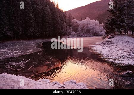 Partially frozen river flowing through snow-covered forest valley at sunset, with ice formations and mountains in the background. Stock Photo