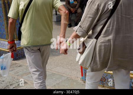 Senior couple holding hands, exploring Thai temple, travelers walking through sacred temple space, rear view. Stock Photo