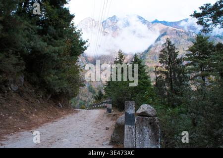 A narrow unpaved dirt road winding through steep rocky cliffs and lush green hills, scenic Marsyangdi river valley view under cloudy sky on the Annapu Stock Photo