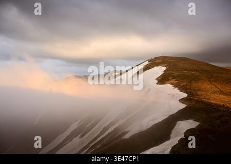 Mountain slope covered with snow streaks and rolling fog under cloudy sky. Misty atmosphere and dramatic terrain create cinematic wilderness mood Stock Photo