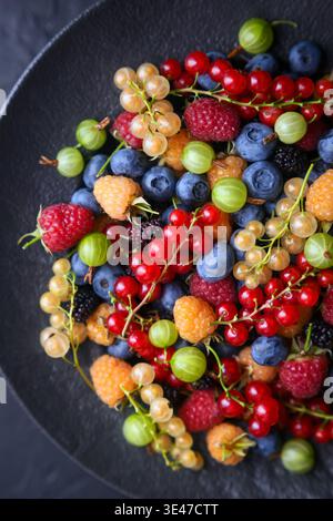 Fresh assorted berries in black ceramic plate, minimal dark styling. Seasonal colorful fruit mix, healthy eating concept Stock Photo