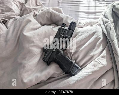 Modern black semi-automatic handgun with tactical flashlight optical red dot sight and extended magazine lying on soft grey bed sheets. Stock Photo