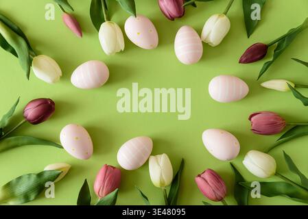 Festive spring flat lay featuring pastel Easter eggs arranged in a circular frame surrounded by white and deep purple tulips on a bright green backgro Stock Photo
