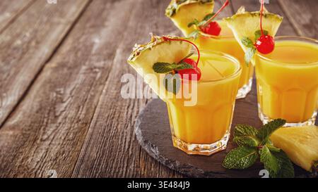 Glasses of pineapple juice with pieces of pineapple, cocktail cherry  and mint on wooden table, healthy drink concept. Stock Photo