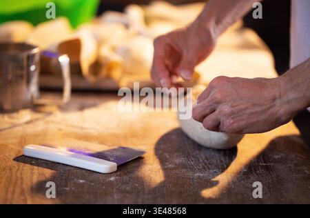 Detailed shot of human hands working with raw dough on rustic wooden surface. Dusting of flour visible, dough scraper tool lying nearby. Focus on hand Stock Photo