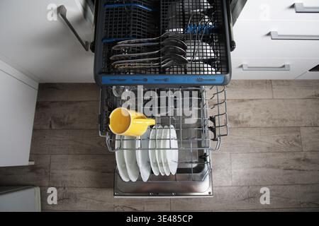 Open modern dishwasher shows clean dishes. Upper rack contains cutlery. Lower rack holds plates, yellow mug, glass. Kitchen setting: white cabinets, w Stock Photo
