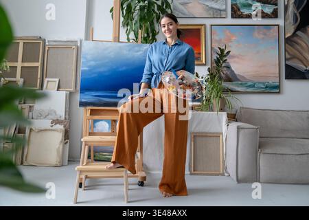 Smiling successful female painter posing in front of artwork on easel in modern art home studio. Stock Photo
