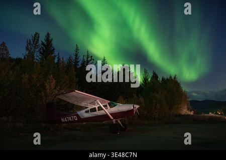 View of a small red and white airplane parked on a dark field beneath the vibrant, swirling green aurora borealis, Coldfoot, Alaska, United States. Stock Photo