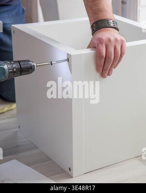 Man assembling white cabinet with cordless drill, driving screw into furniture panel, DIY home improvement, flat pack furniture assembly, handyman Stock Photo
