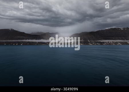 View of rugged dark mountains and deep blue sea under heavy storm clouds with low-lying mist in Greenland. Stock Photo