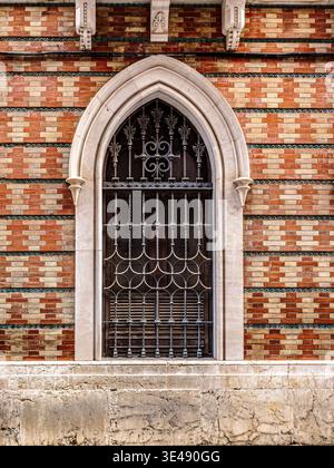Gothic arched window with ornate wrought-iron grille and carved stone surround set in a colorful patterned brick façade, Malaga, Spain Stock Photo