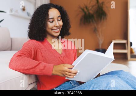 Young Woman Sits on Floor and Reads Notebook While Relaxing at Home in Bright Room Stock Photo