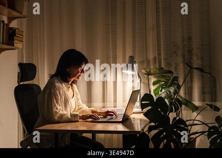 Woman focusing on work while sitting desk with laptop lamp illuminating her home office workspace, working on laptop late night Stock Photo